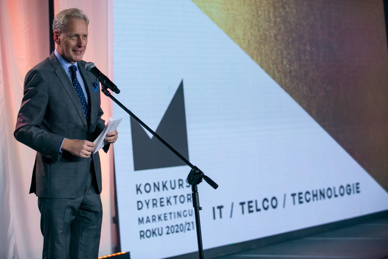 Siwowłosy wręczający w szarym garniturze zapowiada kategorię IT, telco i technologie — Gala DMR X (2021)