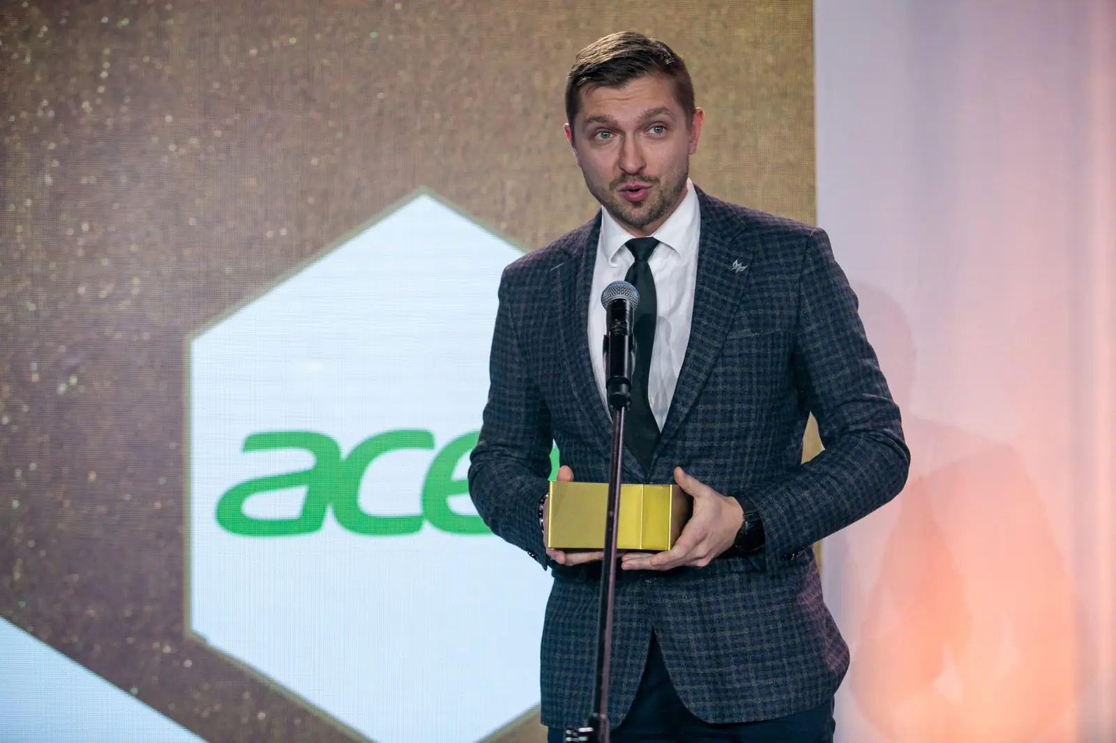 Laureat w szarym garniturze w kratę mówi do mikrofonu ze złotą statuetką przed sobą — Gala DMR X (2021)