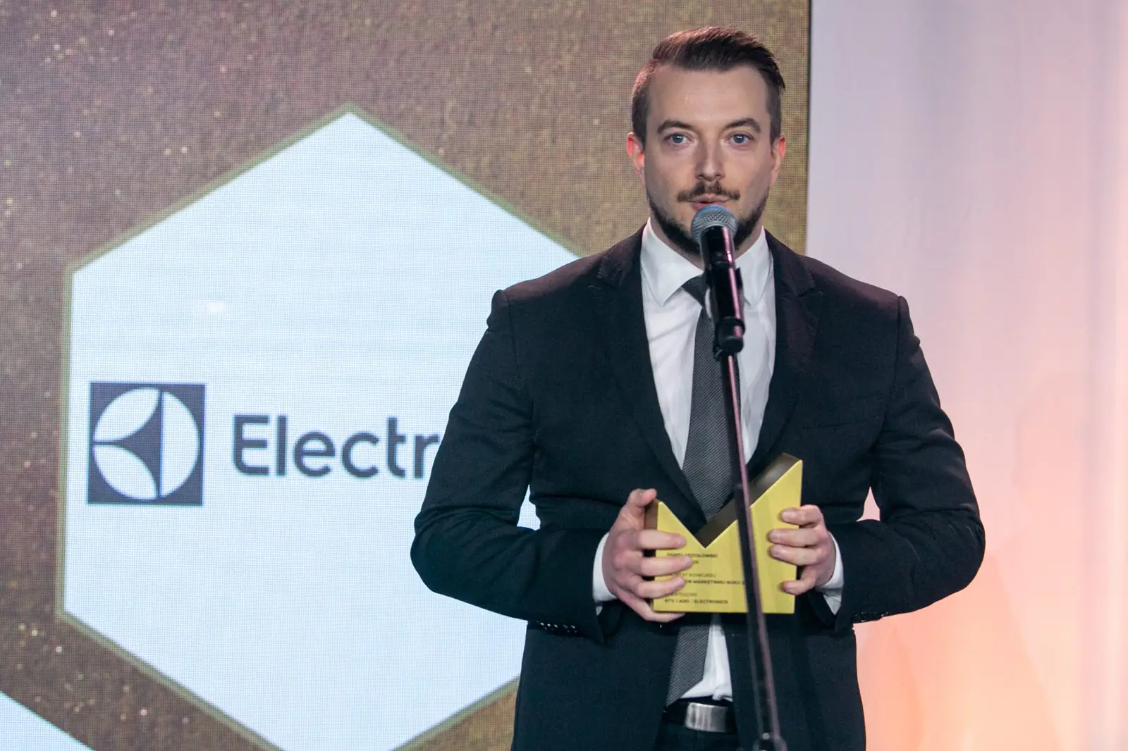 Laureat z wąsem i bródką w czarnym garniturze dziękuje ze statuetką przed logo firmy — Gala DMR X (2021)