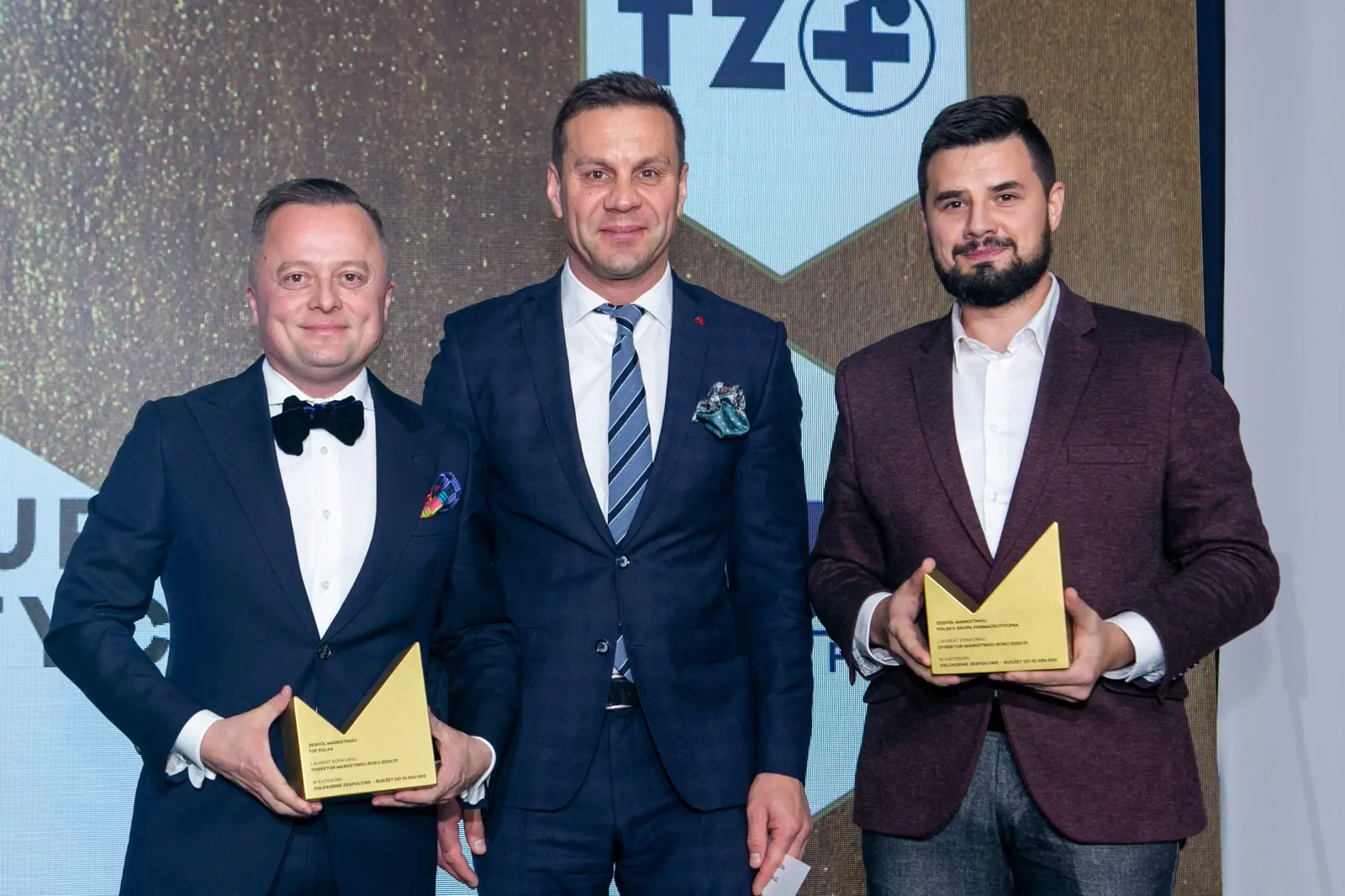 Two winners with statuettes, one in a bow tie and one in a burgundy jacket, pose with the presenter — DMR Gala X (2021)