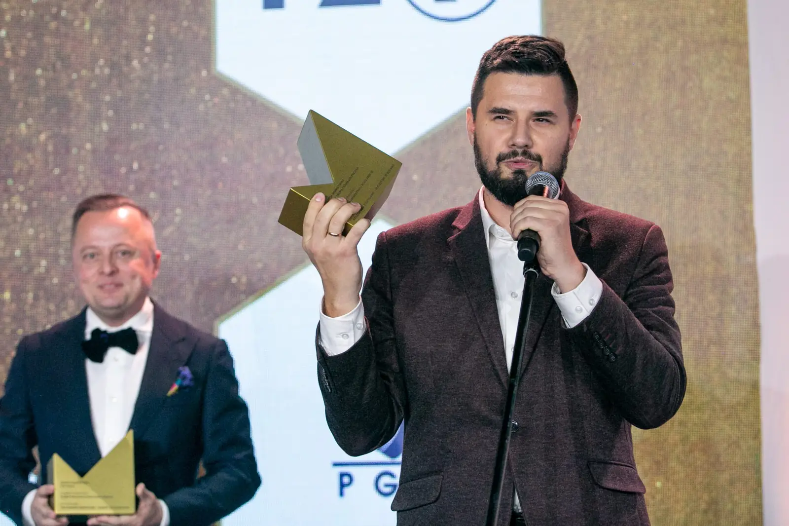 Brodaty laureat unosi statuetkę, mówiąc do mikrofonu, a za nim stoi drugi nagrodzony — Gala DMR X (2021)
