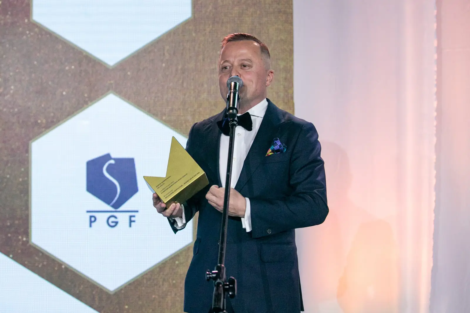 Laureat w muszce i z poszetką dziękuje przy mikrofonie, trzymając złotą statuetkę — Gala DMR X (2021)