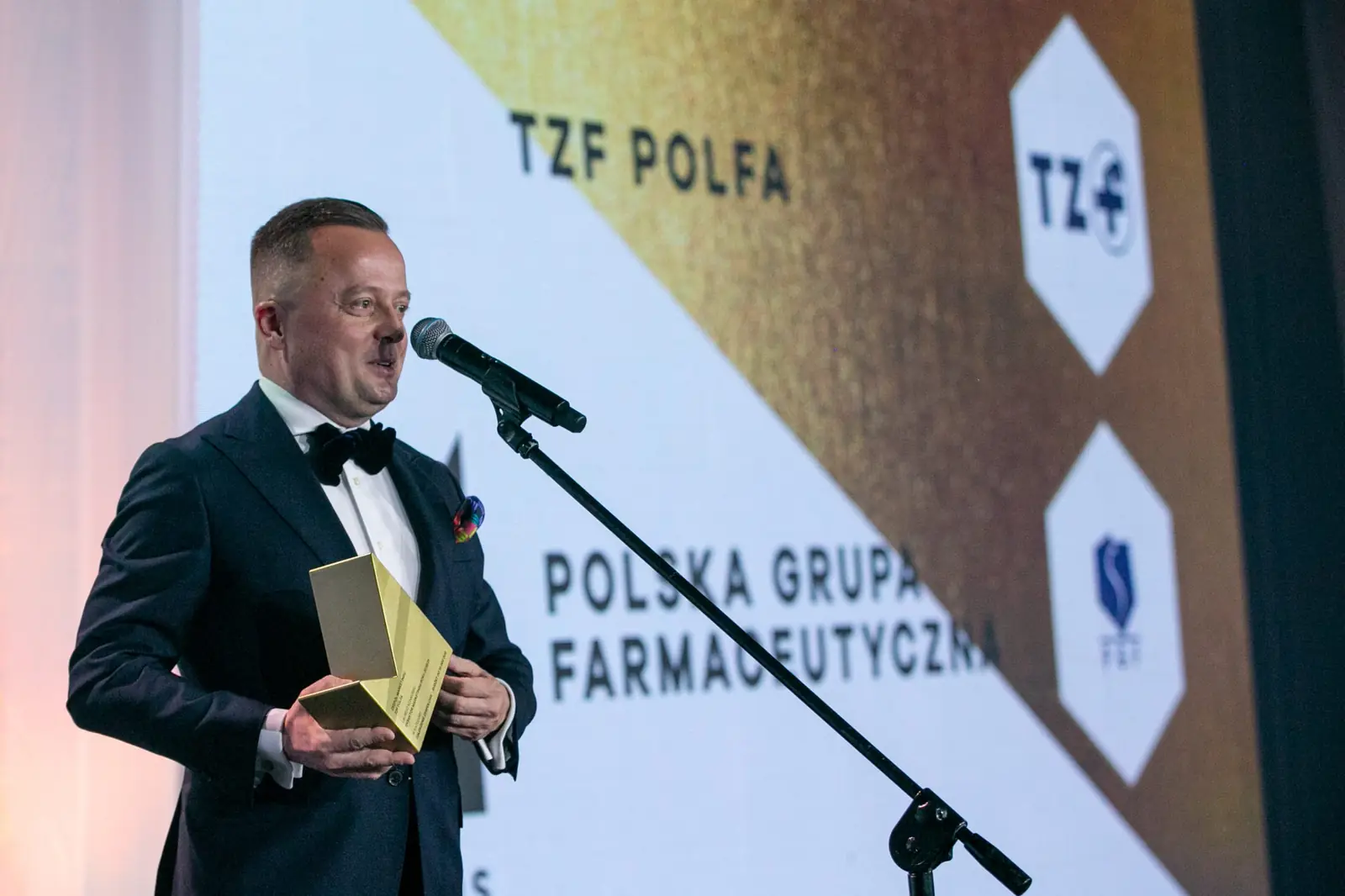 Winner in a bow tie speaks at a stand microphone before a screen showing two company names — DMR Gala X (2021)