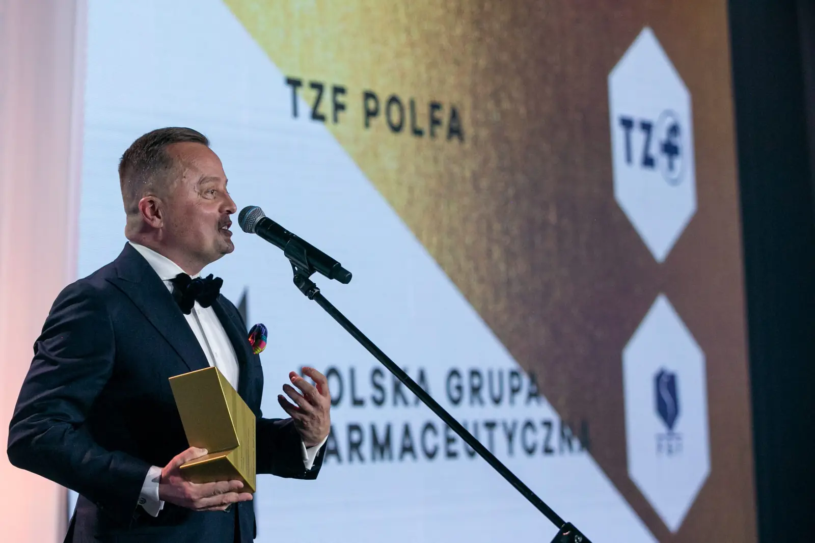 Laureat w muszce przemawia z profilu, trzymając złotą statuetkę przy piersi — Gala DMR X (2021)