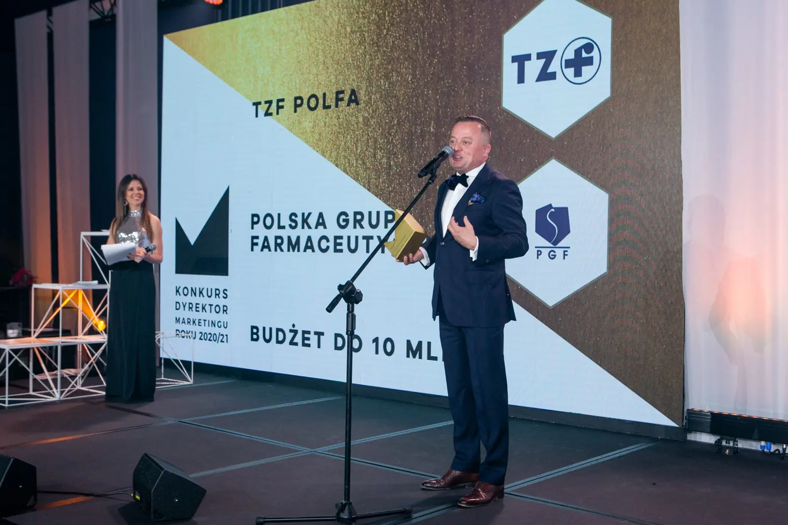 Laureat przemawia na tle logo dwóch nagrodzonych firm, z lewej stoi prowadząca w czarnej sukni — Gala DMR X (2021)