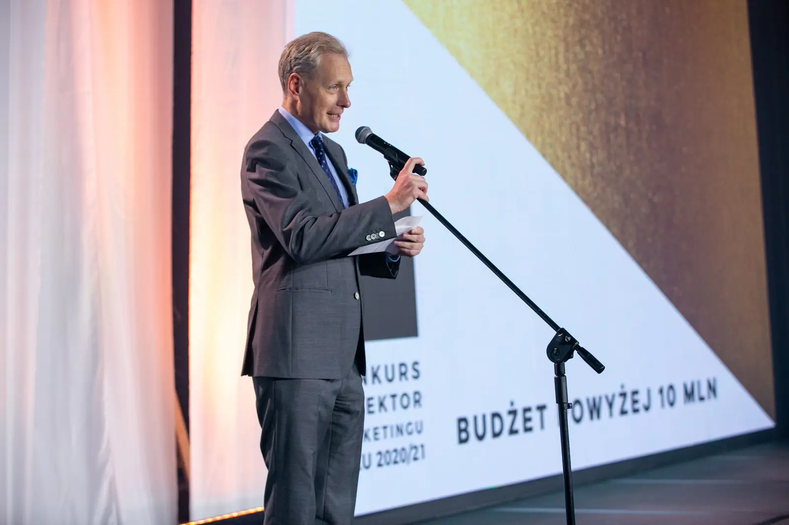 Siwowłosy wręczający zapowiada kategorię budżet powyżej 10 mln, trzymając kartę — Gala DMR X (2021)
