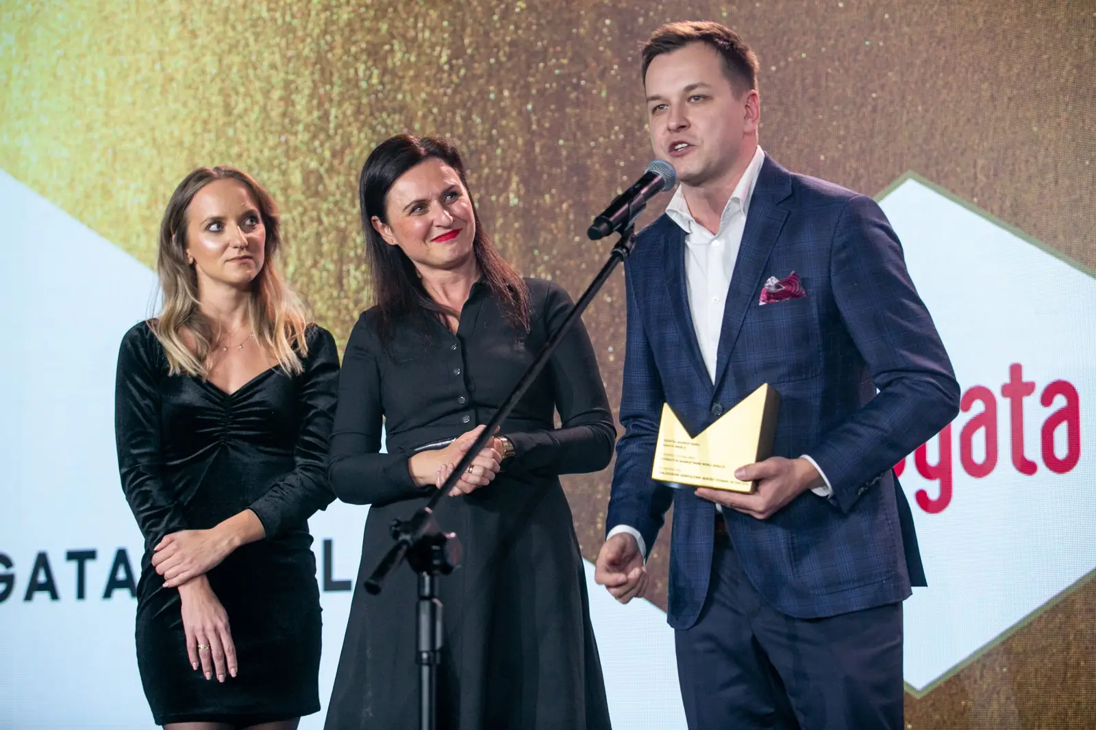 Laureat w granatowym garniturze przemawia, a dwie koleżanki z zespołu stoją obok — Gala DMR X (2021)