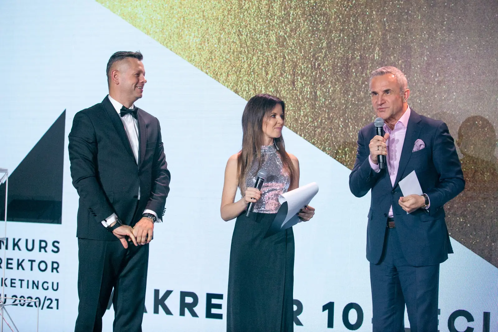 Prowadzący, prowadząca ze szklaną statuetką i siwowłosy mężczyzna z mikrofonem na scenie — Gala DMR X (2021)
