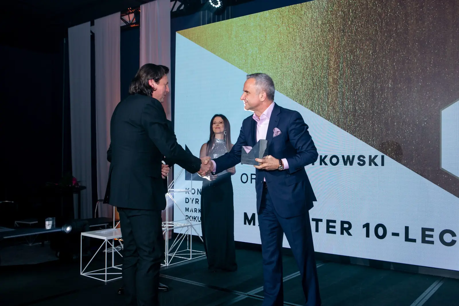 Laureat tytułu Marketer 10-lecia ściska dłoń siwowłosego wręczającego ze statuetką — Gala DMR X (2021)