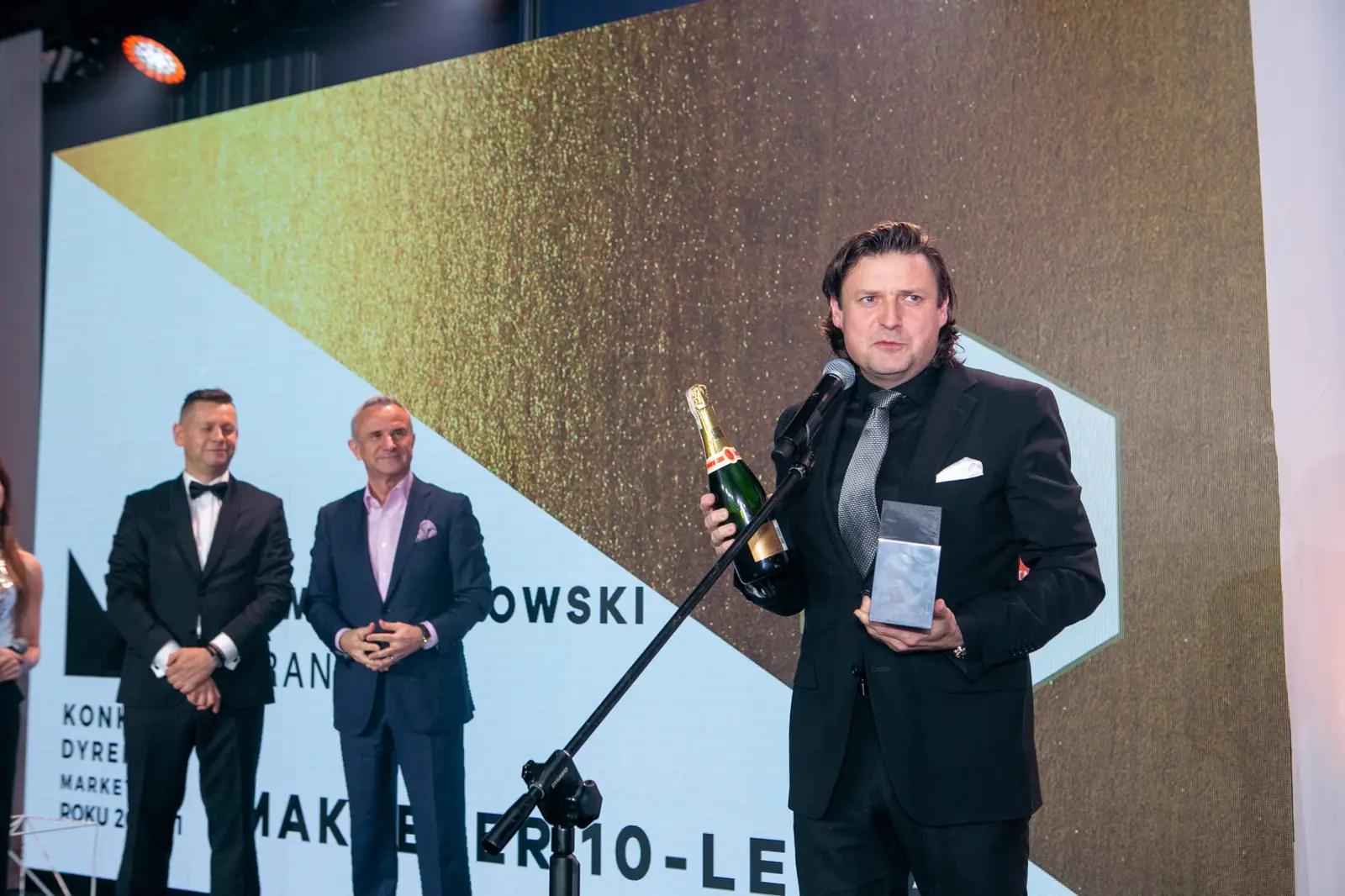 Marketer 10-lecia z butelką szampana i szklaną statuetką przemawia przy mikrofonie — Gala DMR X (2021)