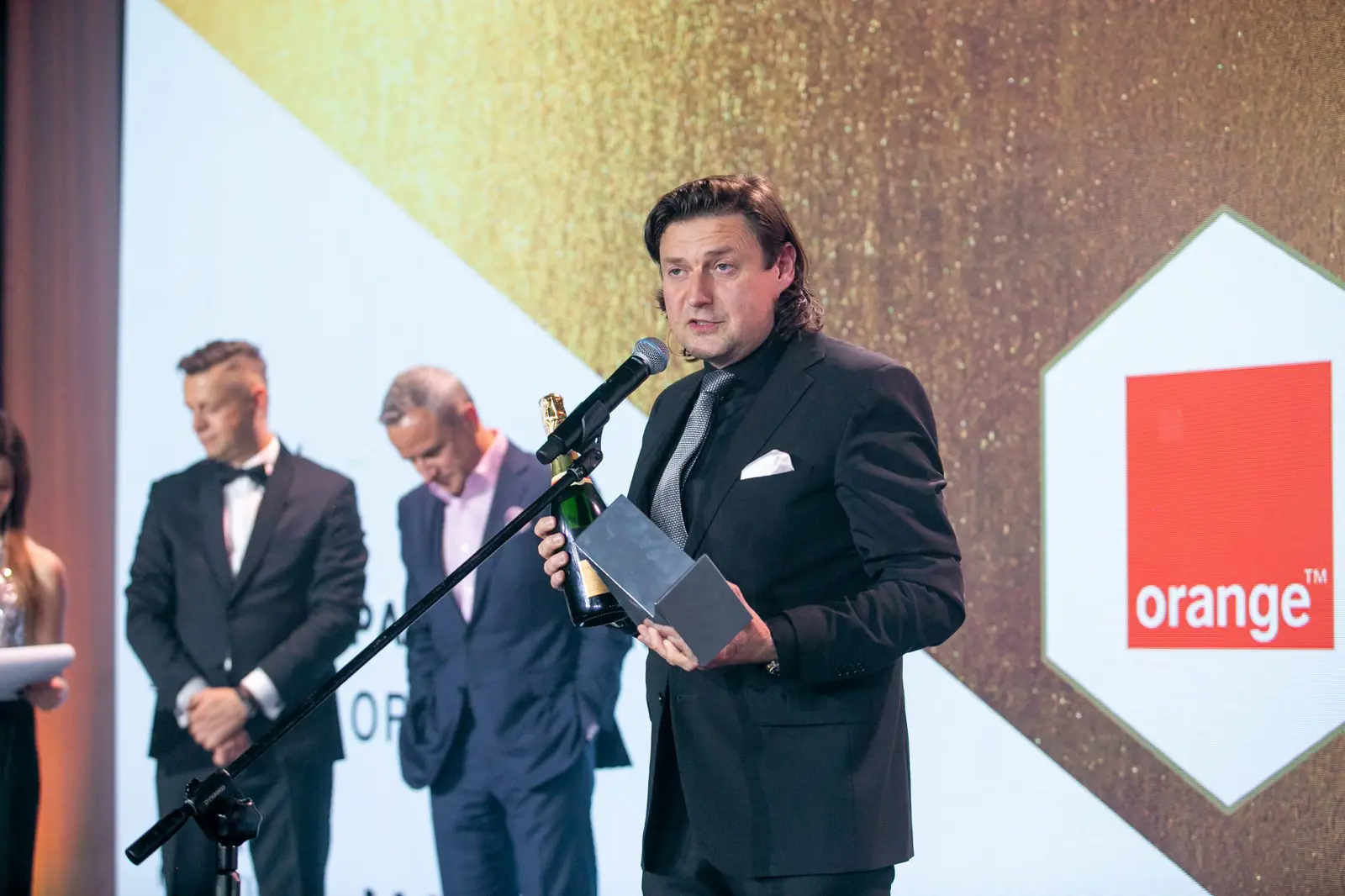 Laureat z długimi włosami mówi do mikrofonu, a prowadzący i wręczający słuchają za nim — Gala DMR X (2021)