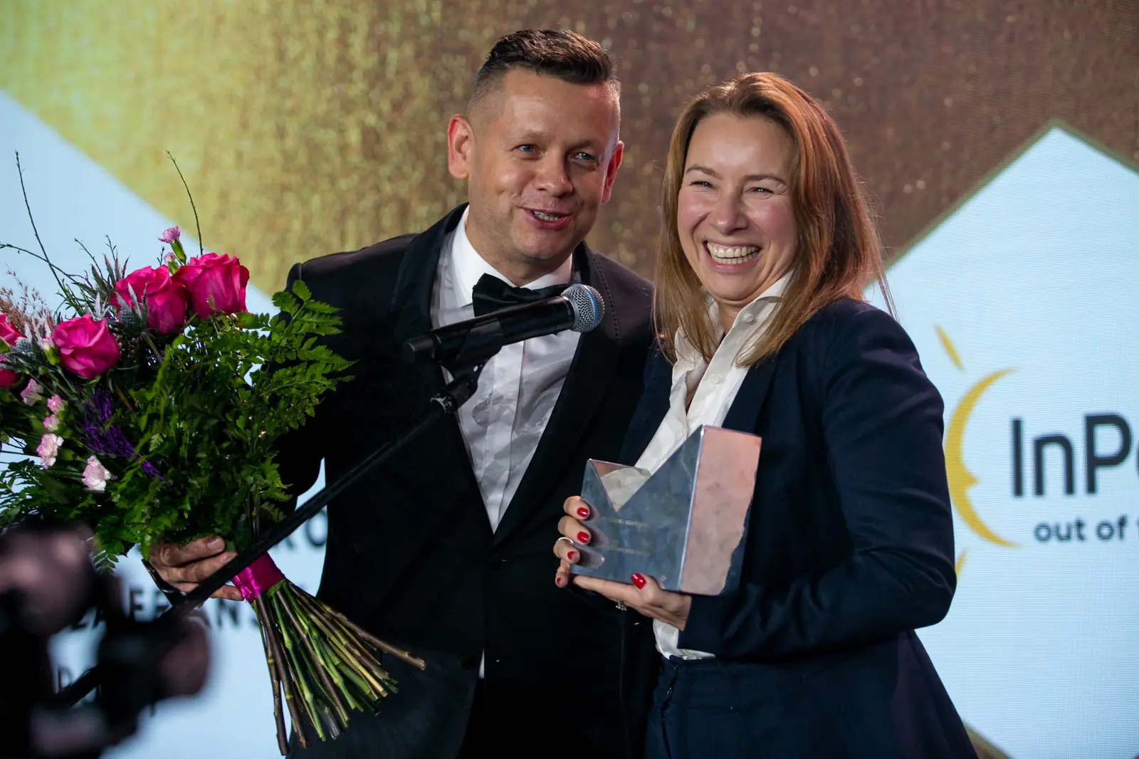 Laughing Grand Prix winner with her glass statuette beside the host holding a bouquet of roses — DMR Gala X (2021)