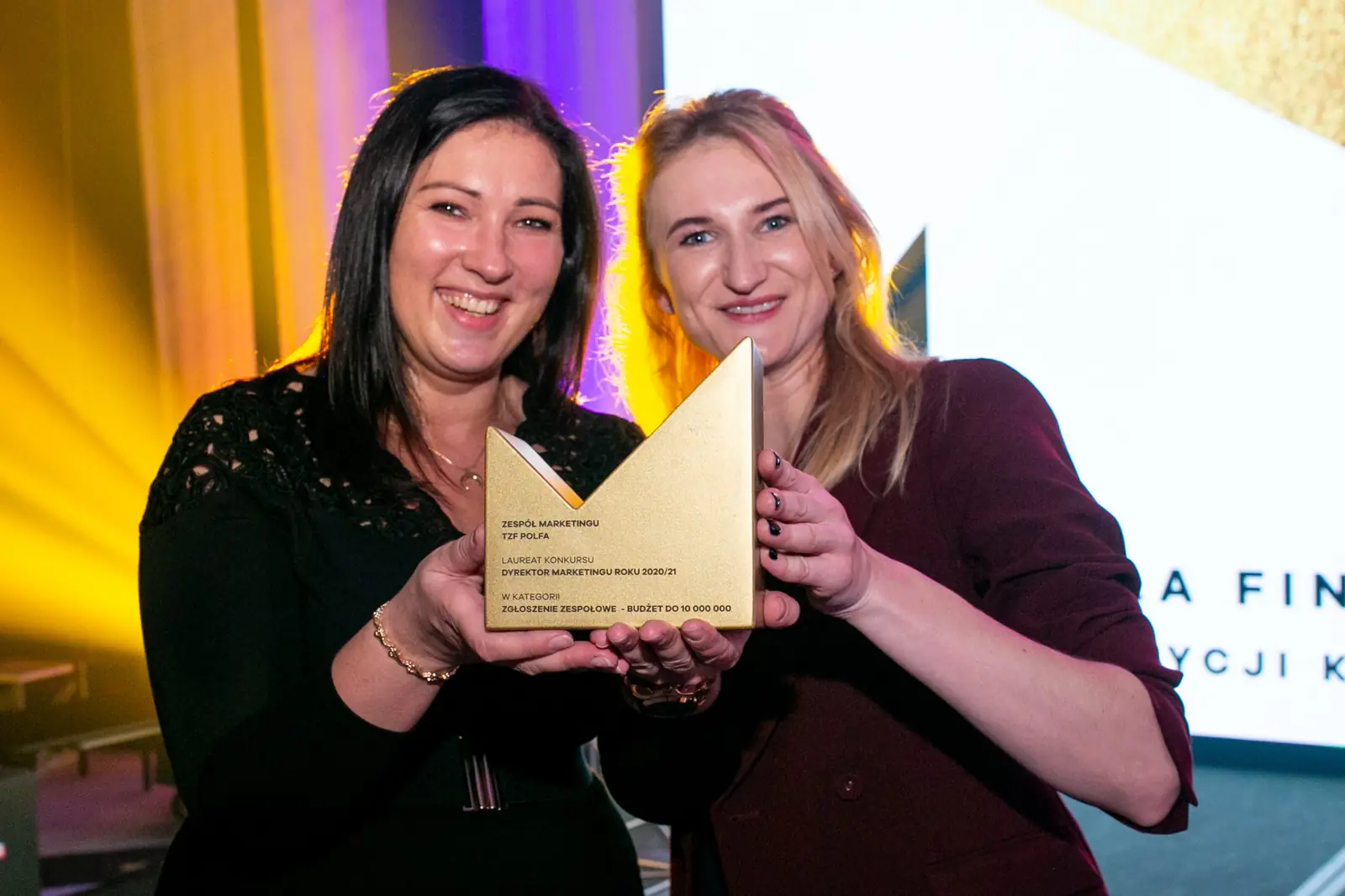 Two smiling women hold a golden statuette together in yellow spotlight — DMR Gala X (2021)