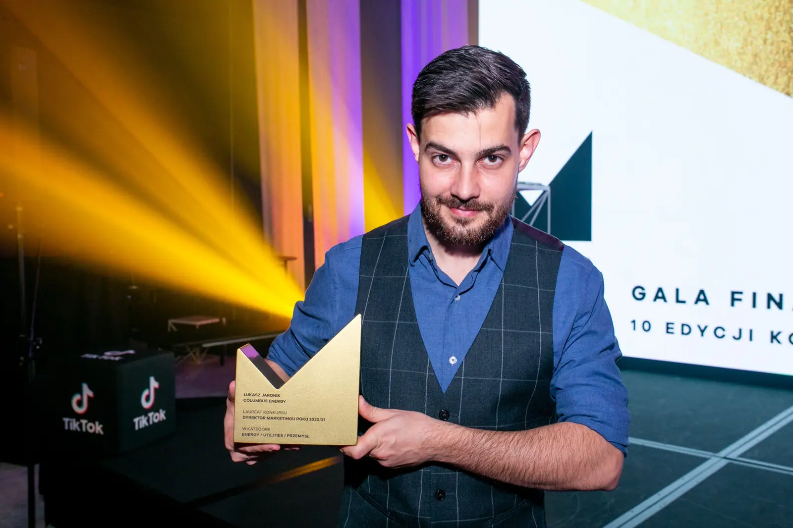 Winner in a waistcoat poses with his statuette against yellow light beams and the gala lettering — DMR Gala X (2021)