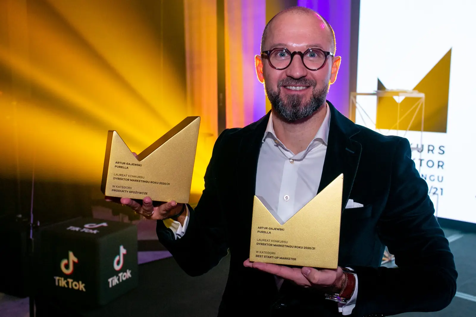Laureat w okularach prezentuje dwie złote statuetki na tle żółtych wiązek światła — Gala DMR X (2021)