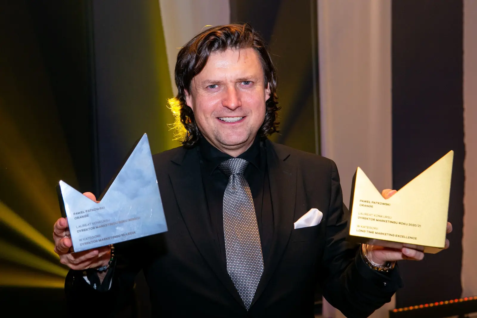 Laureat z długimi włosami pozuje ze szklaną i złotą statuetką w obu dłoniach — Gala DMR X (2021)