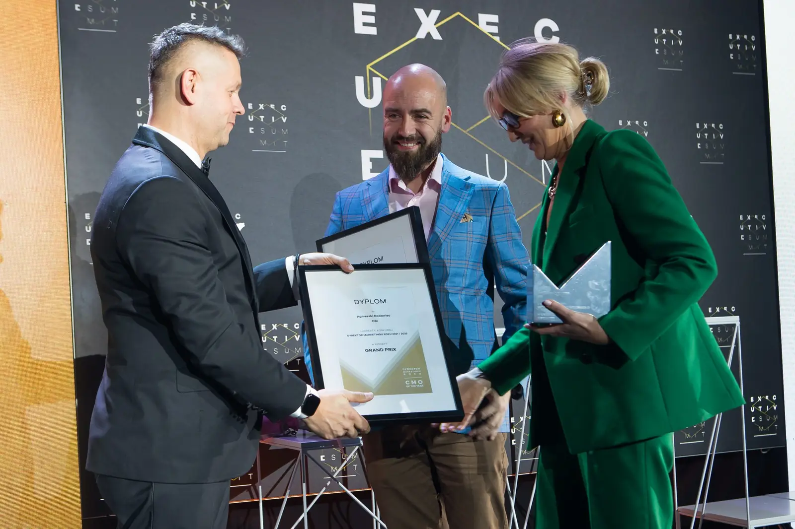 Prowadzący wręcza dyplom Grand Prix laureatowi w niebieskiej marynarce, obok laureatka w zieleni — Gala DMR XI (2022)