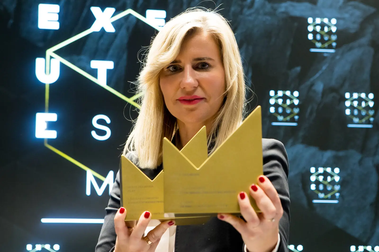 Blondynka z czerwonym manicure trzyma przy twarzy dwie złote statuetki w kształcie litery M — Gala DMR XI (2022)