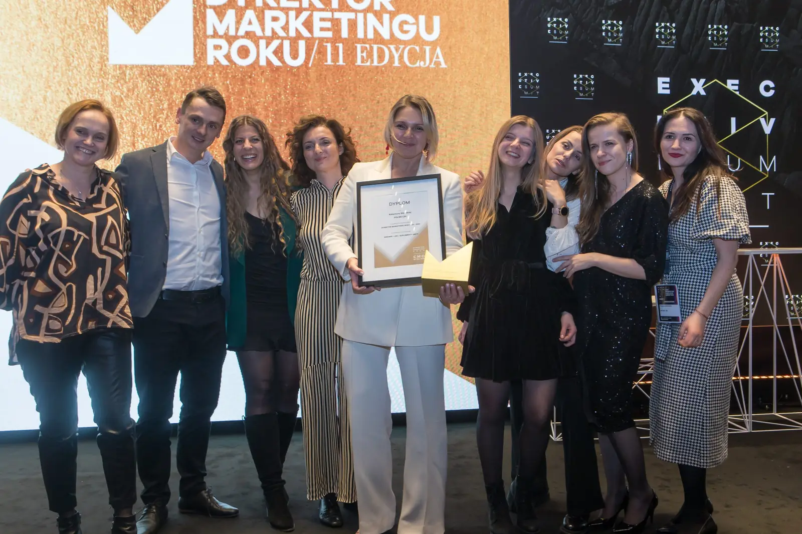Duża grupa współpracowników otacza laureatkę w białym garniturze z dyplomem i statuetką — Gala DMR XI (2022)