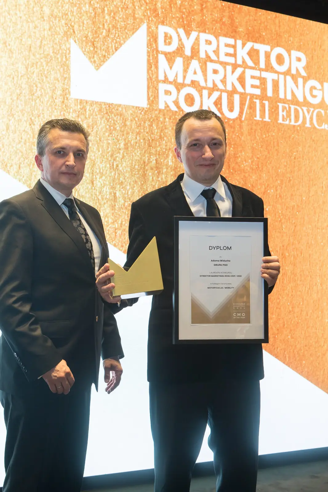 Dwóch mężczyzn w garniturach pozuje ze statuetką i dyplomem przed złotym logo konkursu — Gala DMR XI (2022)