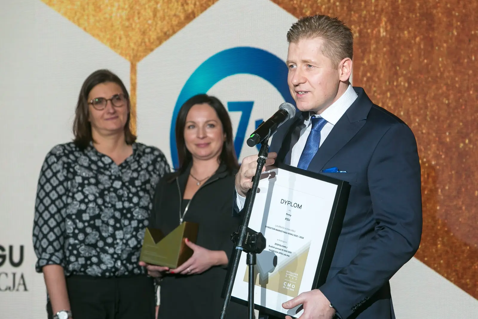 Laureat w granatowym garniturze przemawia przy mikrofonie na statywie, trzymając dyplom — Gala DMR XI (2022)