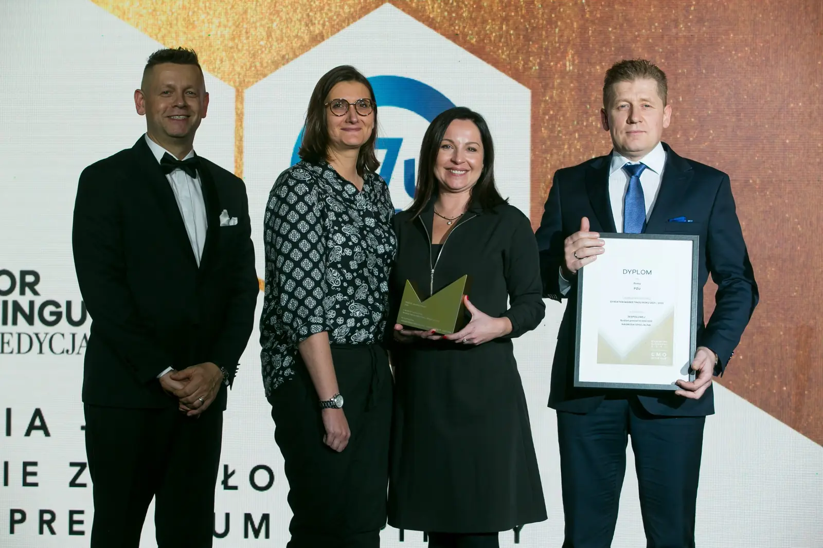 Prowadzący w smokingu stoi obok trojga laureatów trzymających statuetkę i dyplom — Gala DMR XI (2022)