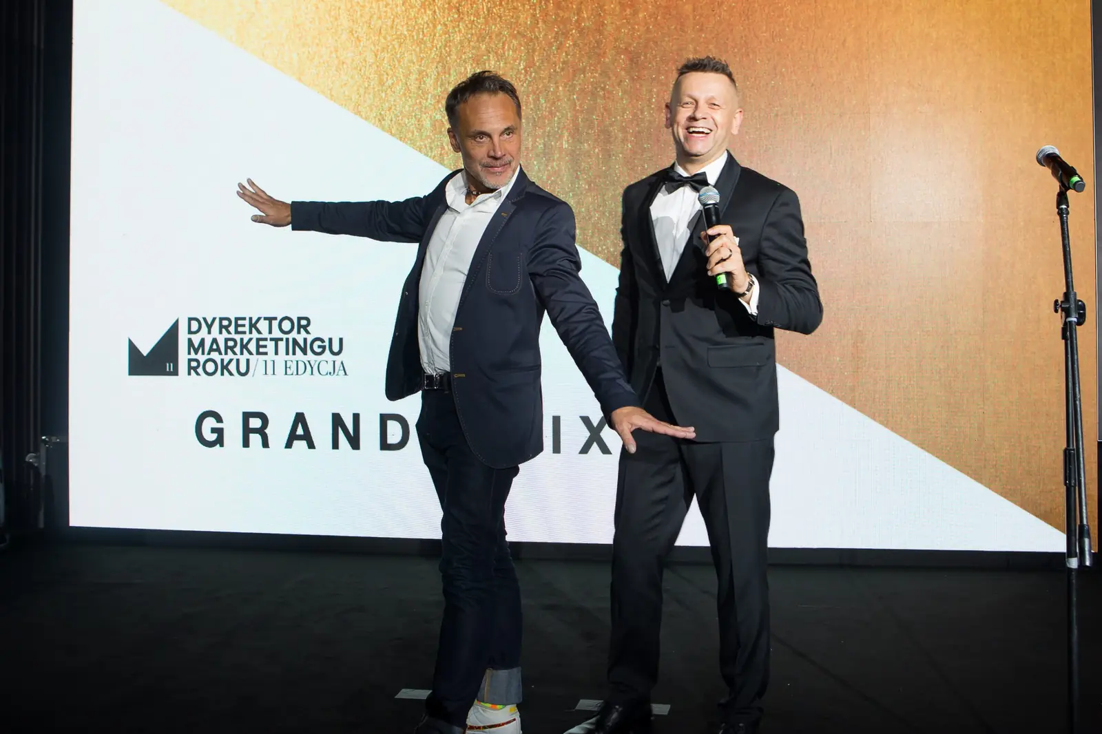 Dwaj prowadzący zapowiadają Grand Prix, jeden z nich wskazuje dłonią ekran z logo — Gala DMR XI (2022)
