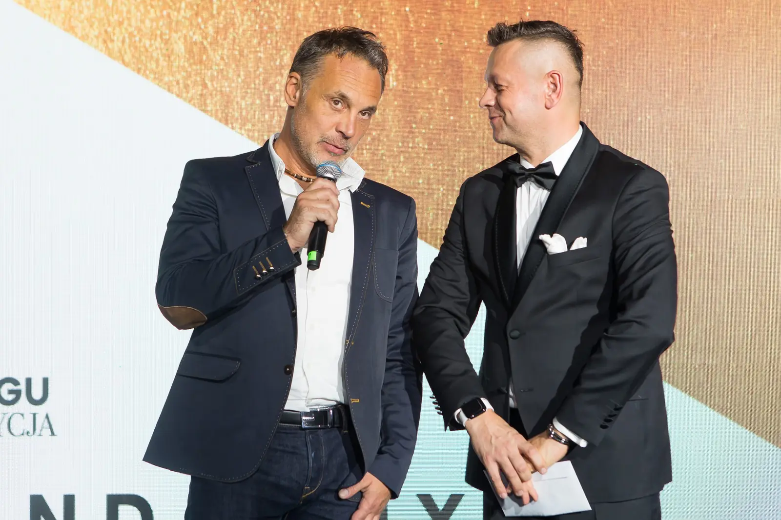Współprowadzący w ciemnej marynarce mówi do mikrofonu, a prowadzący w smokingu słucha — Gala DMR XI (2022)