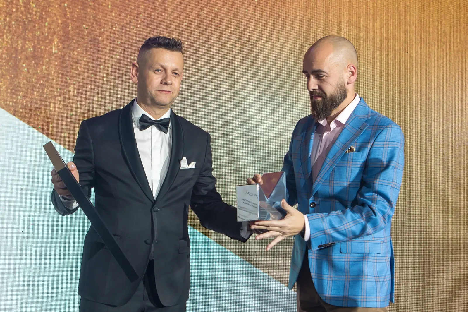 Prowadzący w smokingu wręcza szklaną statuetkę Grand Prix laureatowi w niebieskiej marynarce — Gala DMR XI (2022)