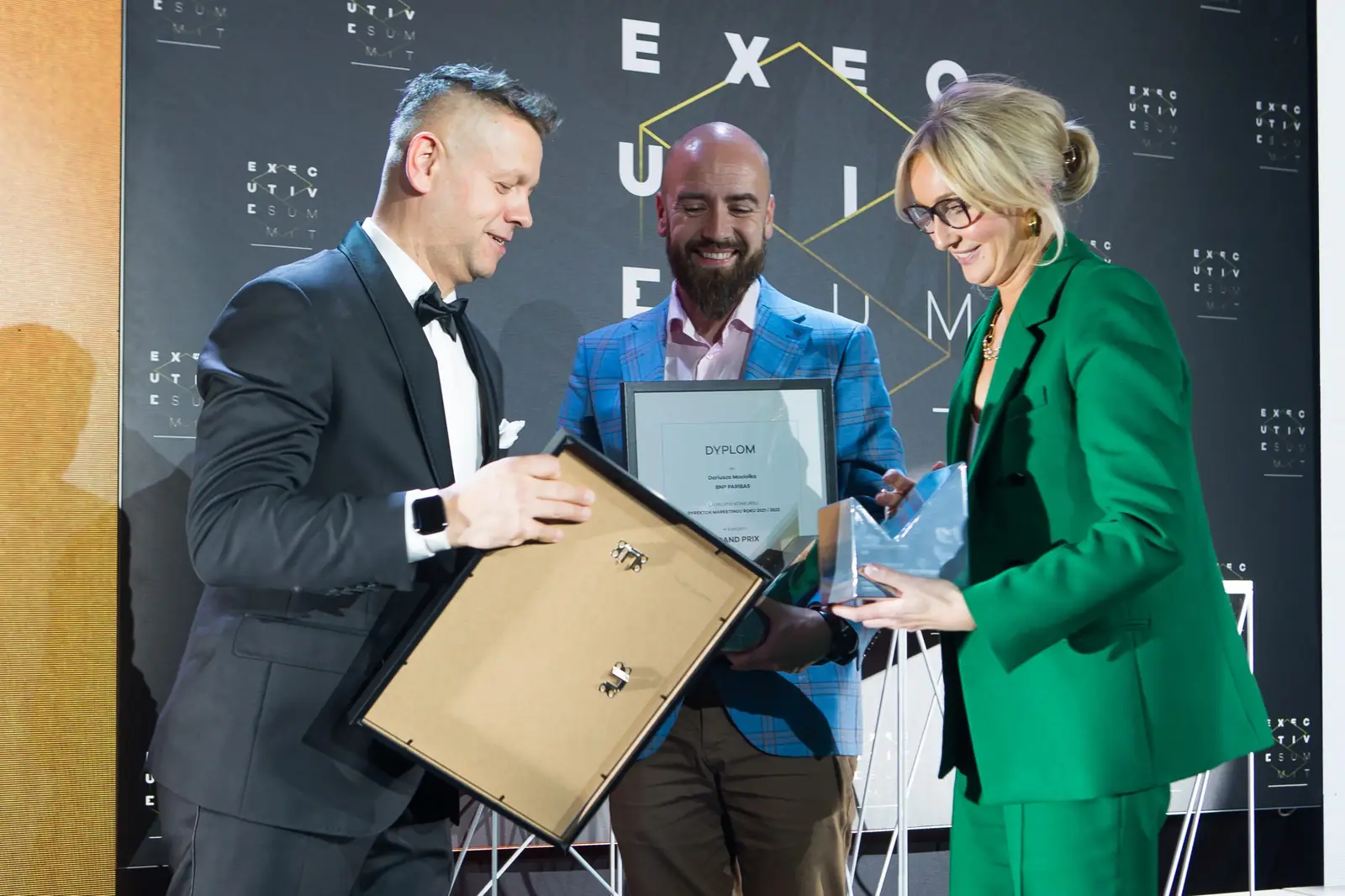 Prowadzący trzyma ramkę dyplomu, a kobieta w zieleni gratuluje laureatowi w niebieskiej marynarce — Gala DMR XI (2022)