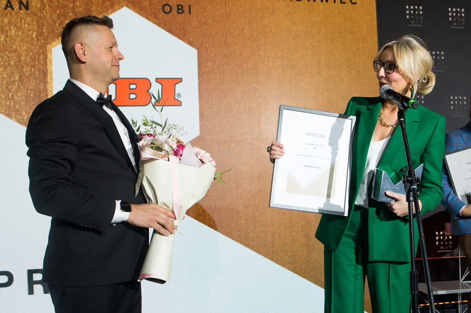 Prowadzący z bukietem kwiatów słucha podziękowań laureatki w zielonym garniturze — Gala DMR XI (2022)