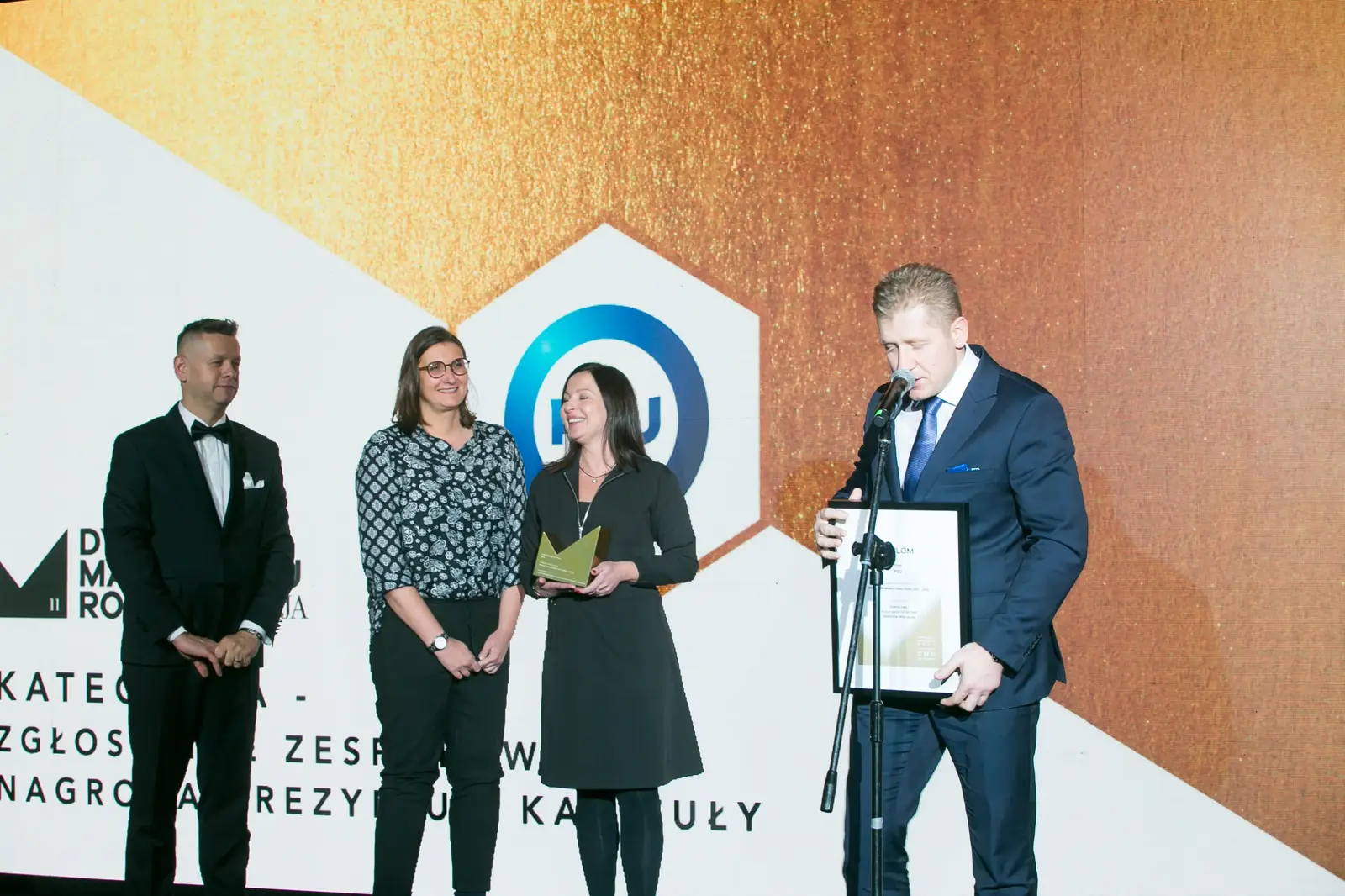 Laureat z dyplomem przemawia przy mikrofonie, obok dwie laureatki i prowadzący w smokingu — Gala DMR XI (2022)