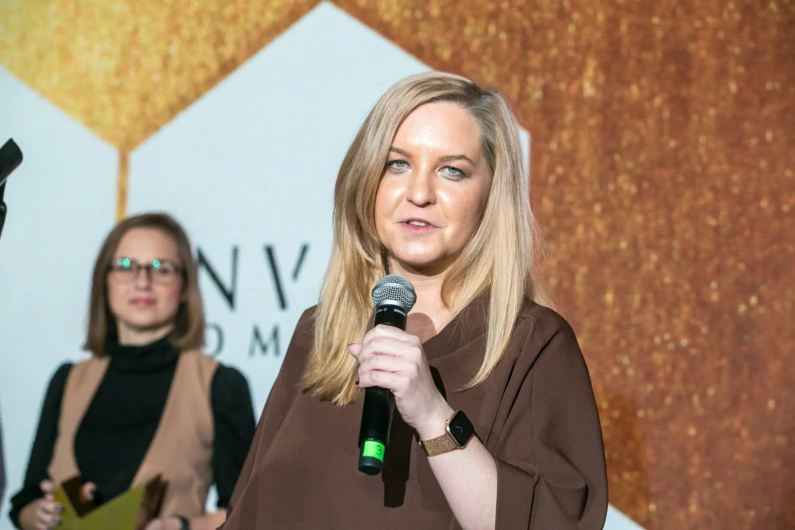 Blondynka w brązowej bluzce przemawia do mikrofonu, za nią stoi laureatka w okularach — Gala DMR XI (2022)