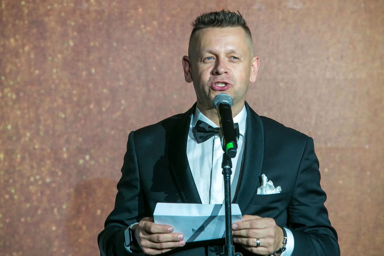 Prowadzący w smokingu z muszką czyta z karty przy mikrofonie na statywie — Gala DMR XI (2022)