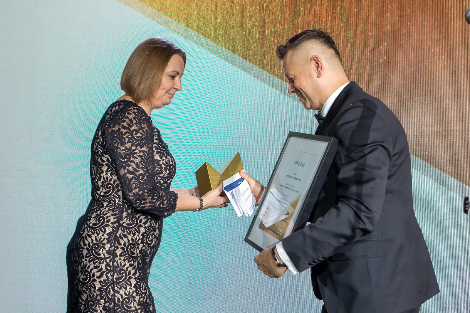 Prowadzący z lekkim ukłonem wręcza oprawiony dyplom laureatce w koronkowej sukience — Gala DMR XI (2022)