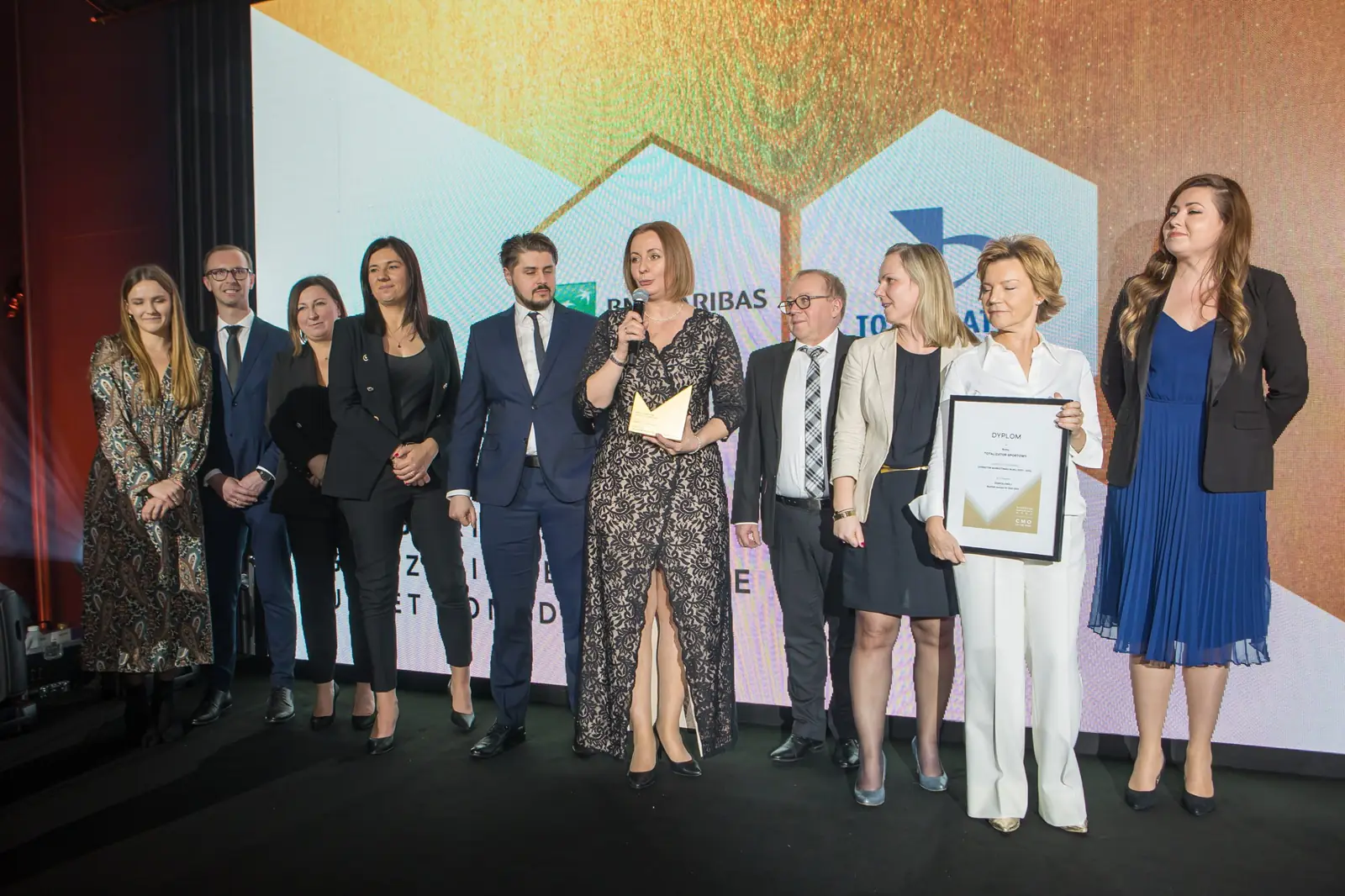 Laureatka w koronkowej sukience ze statuetką pośrodku dużej grupy nagrodzonych na scenie — Gala DMR XI (2022)