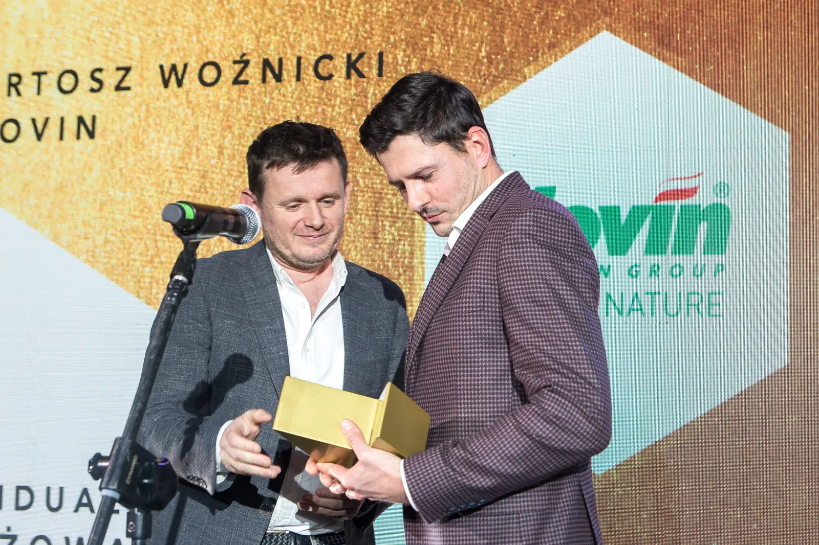 Wręczający w szarej marynarce podaje złotą statuetkę laureatowi w marynarce w kratę — Gala DMR XI (2022)
