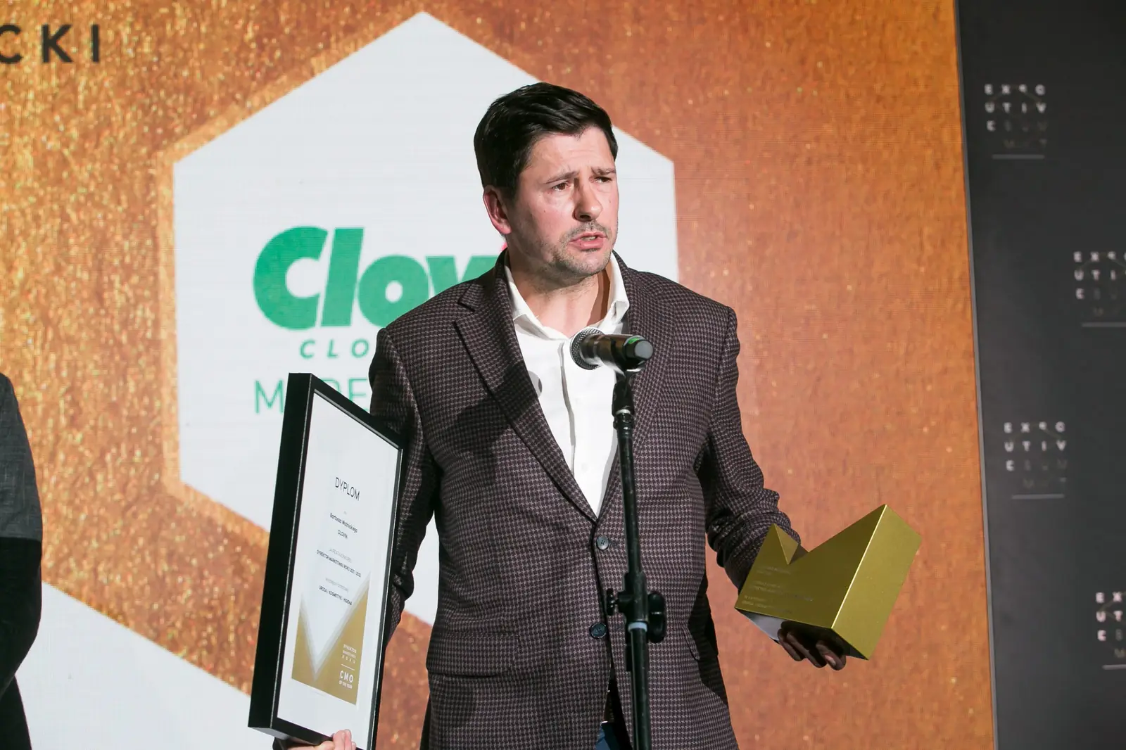 Laureat w brązowej marynarce w kratę przemawia przy mikrofonie z dyplomem i statuetką — Gala DMR XI (2022)