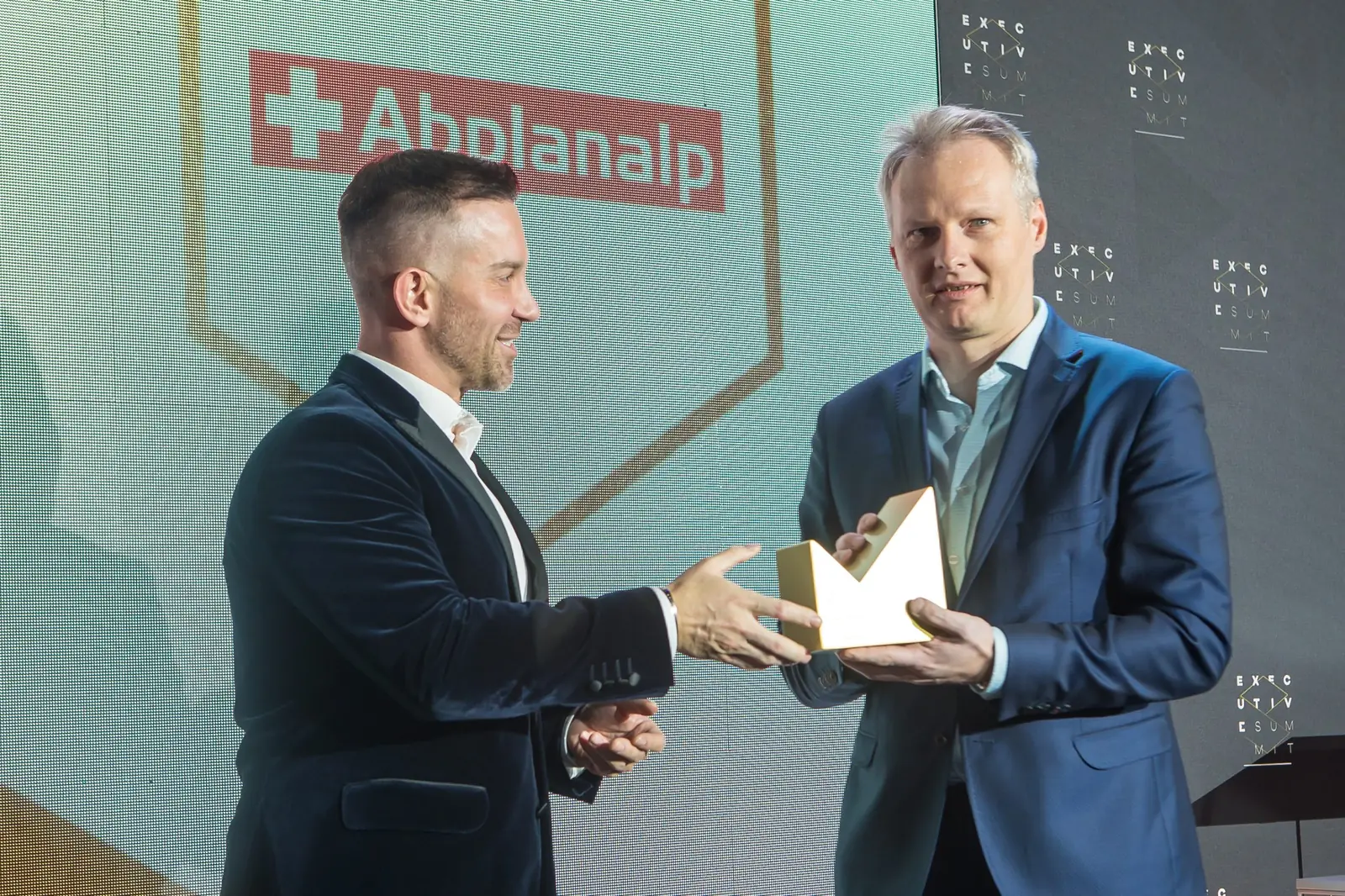 Mężczyzna w aksamitnej marynarce wręcza złotą statuetkę laureatowi w granatowym garniturze — Gala DMR XI (2022)