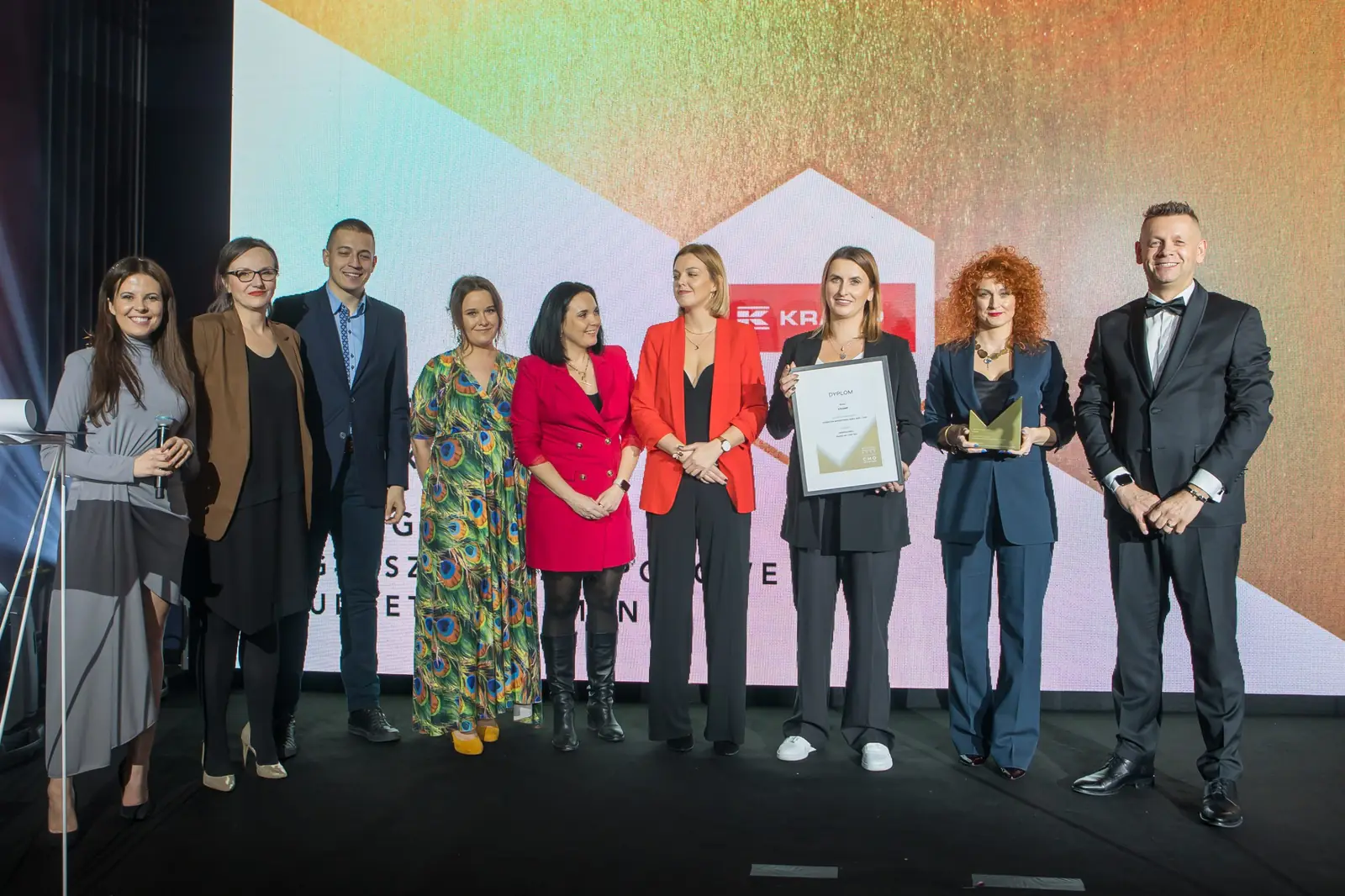 Dziesięcioosobowy zespół z rudowłosą laureatką pozuje na scenie wraz z prowadzącym — Gala DMR XI (2022)