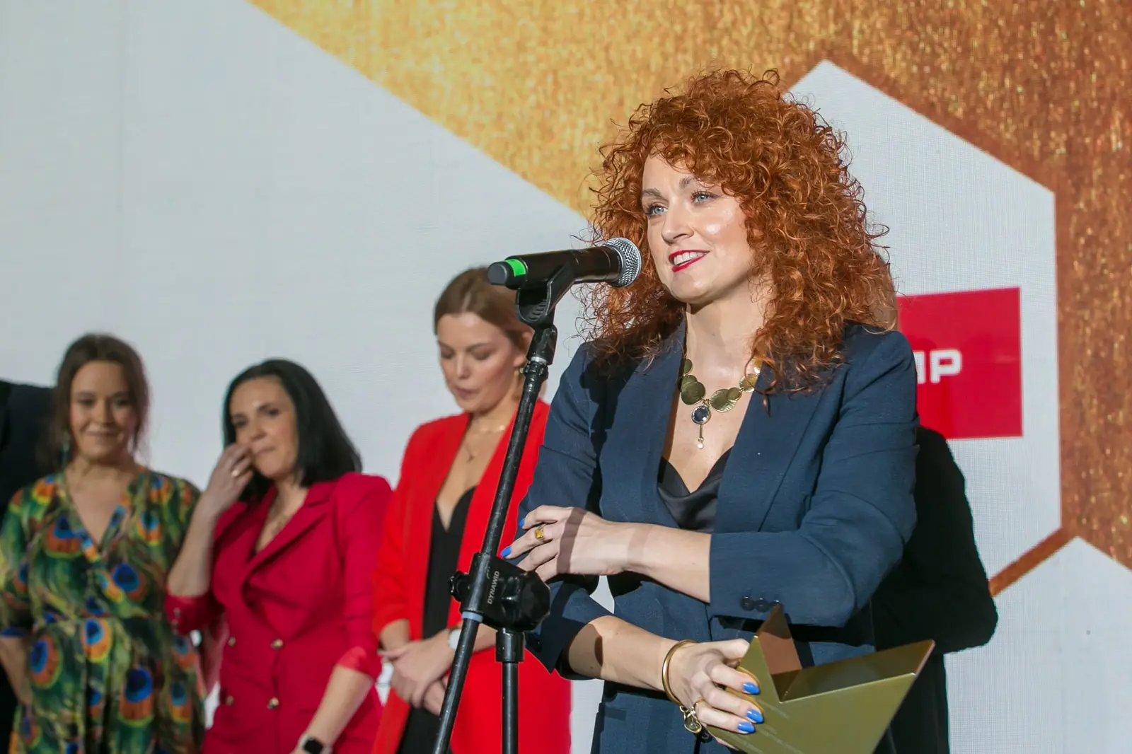 Laureatka z rudymi lokami spogląda na statuetkę, a koleżanki w czerwieni słuchają jej słów — Gala DMR XI (2022)