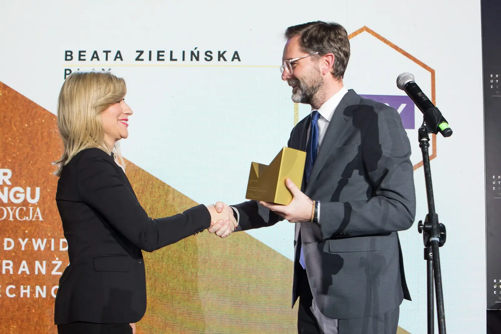 Wręczający w okularach ze złotą statuetką w dłoni ściska rękę laureatki w czarnym żakiecie — Gala DMR XI (2022)