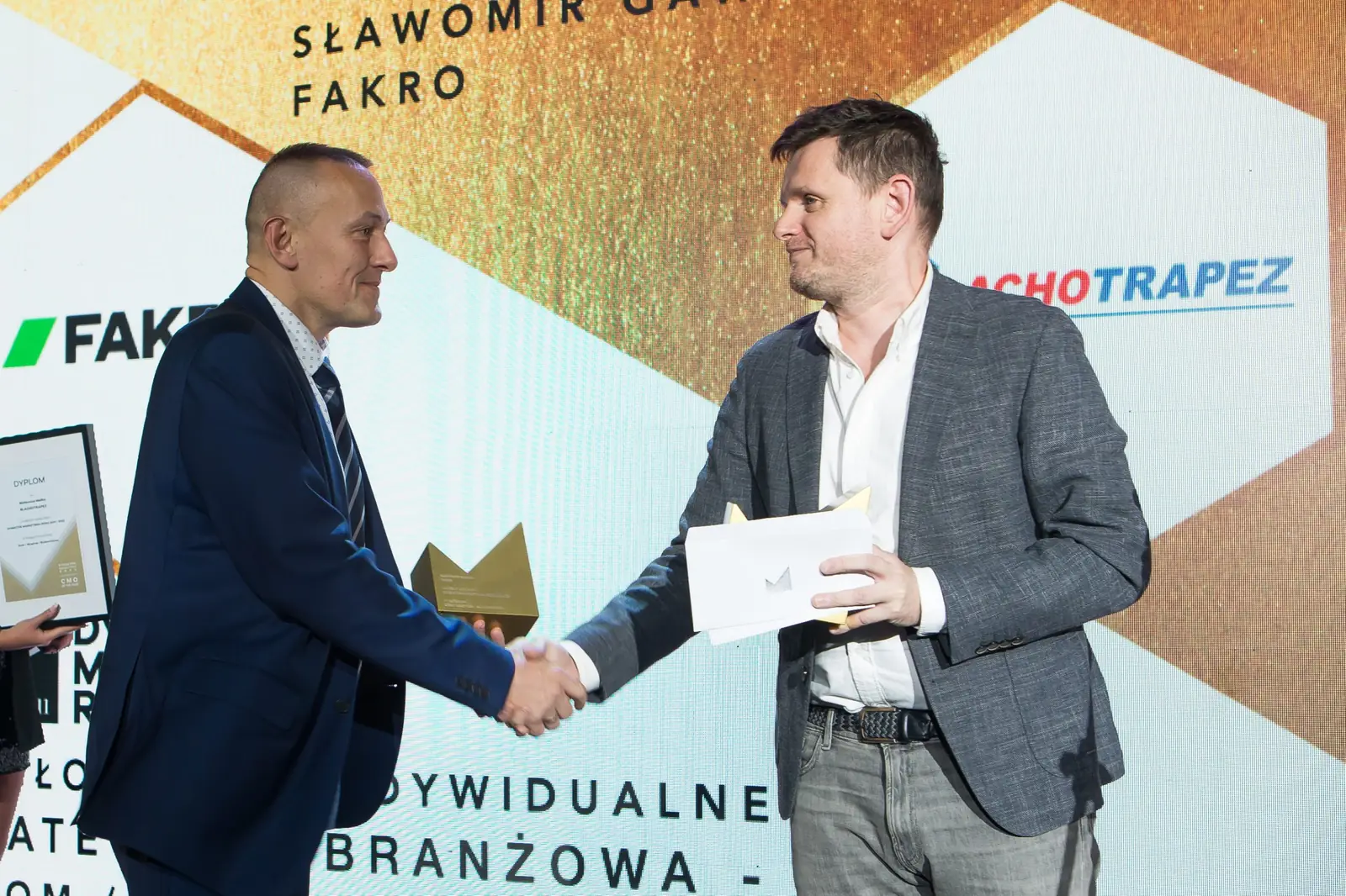 Laureat w granatowym garniturze ściska dłoń wręczającego, trzymając złotą statuetkę — Gala DMR XI (2022)