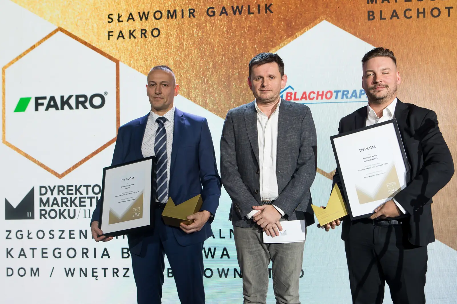 Dwóch laureatów z dyplomami i statuetkami pozuje z wręczającym w szarej marynarce — Gala DMR XI (2022)