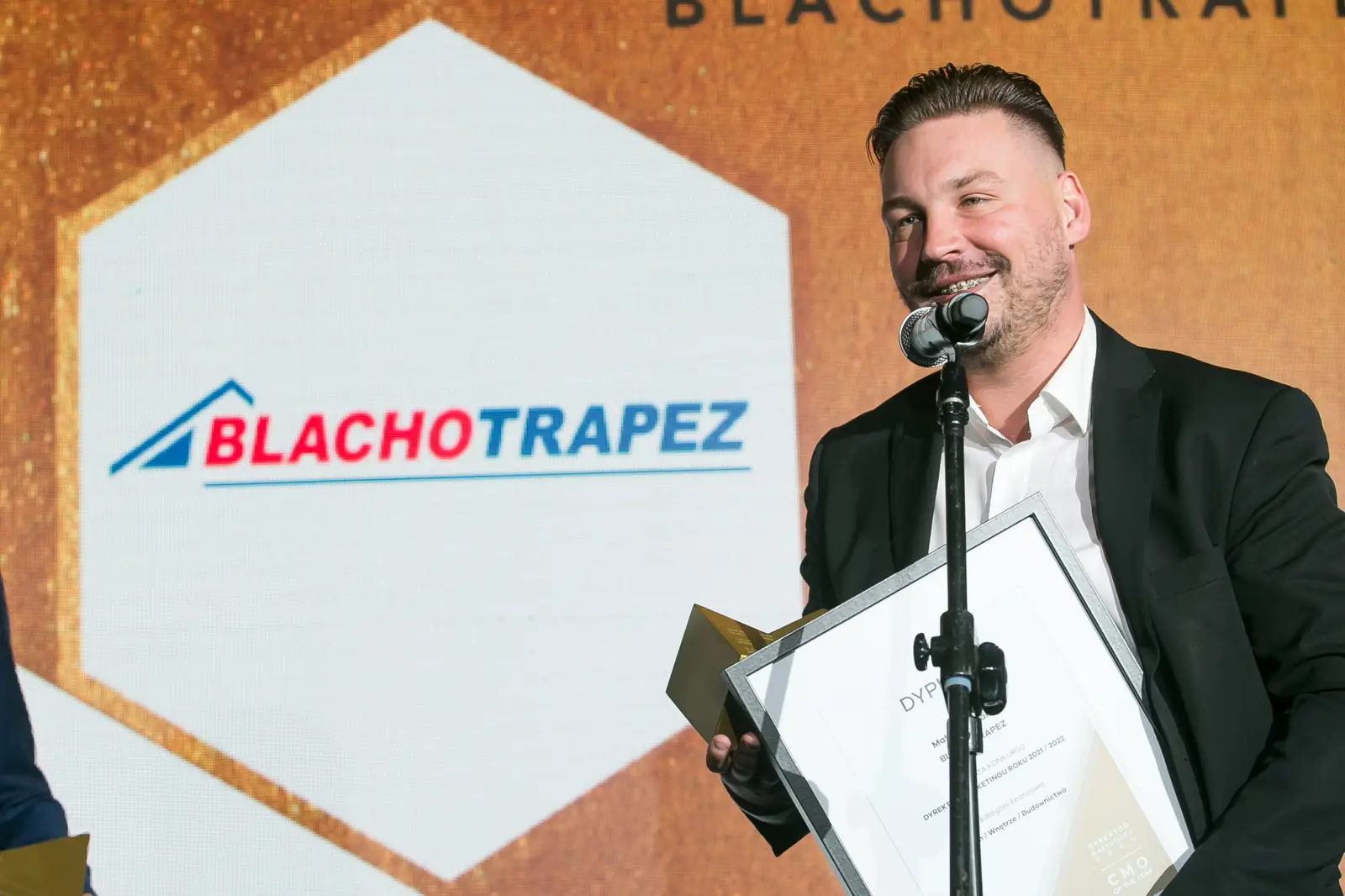 Uśmiechnięty laureat z zarostem przemawia przy mikrofonie z oprawionym dyplomem w dłoni — Gala DMR XI (2022)