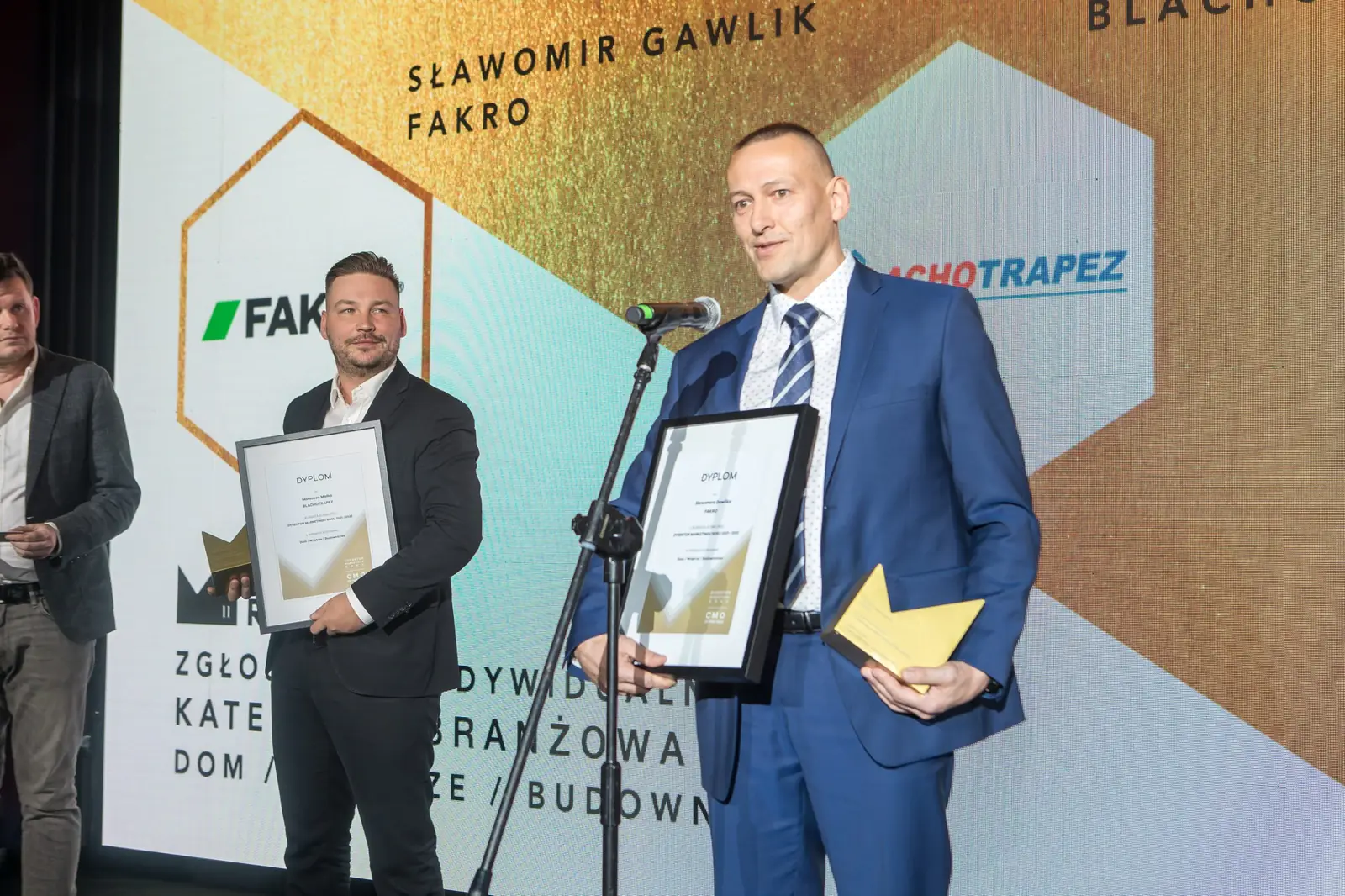 Laureat w granatowym garniturze przemawia z dyplomem i statuetką, za nim drugi nagrodzony — Gala DMR XI (2022)