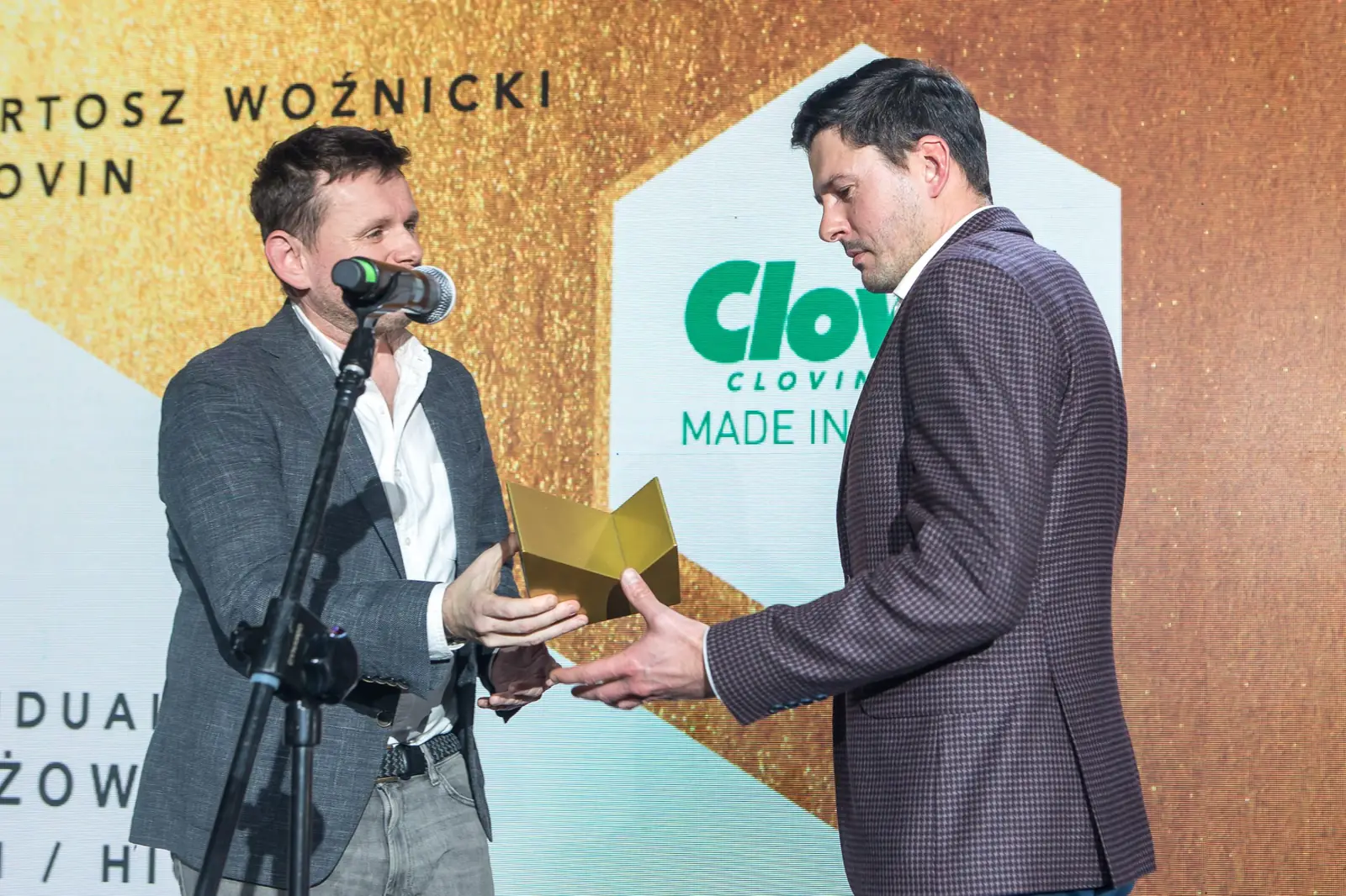 Laureat w marynarce w brązową kratę odbiera statuetkę od wręczającego stojącego przy mikrofonie — Gala DMR XI (2022)