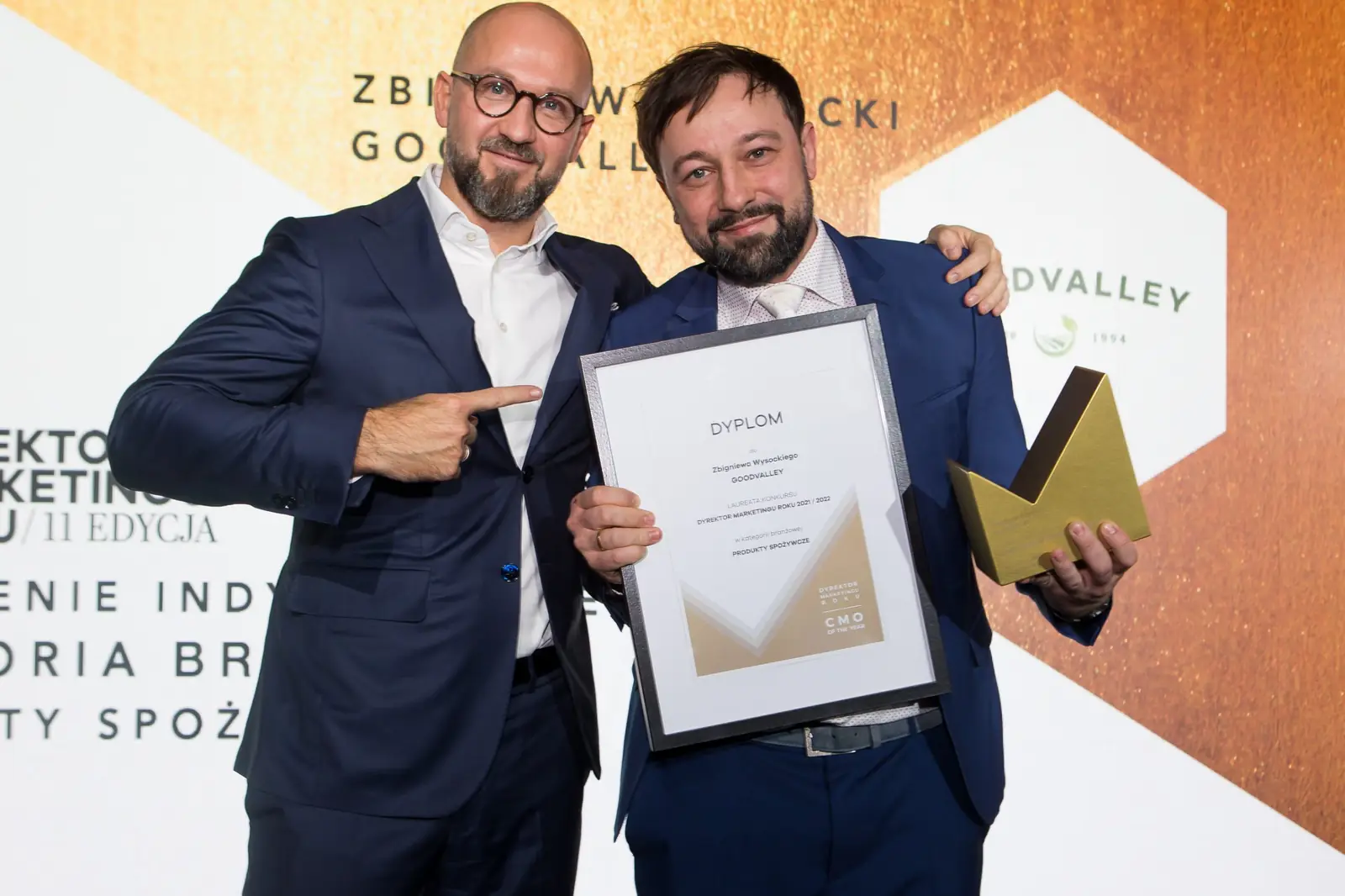Wręczający żartobliwie wskazuje palcem laureata z dyplomem i złotą statuetką — Gala DMR XI (2022)
