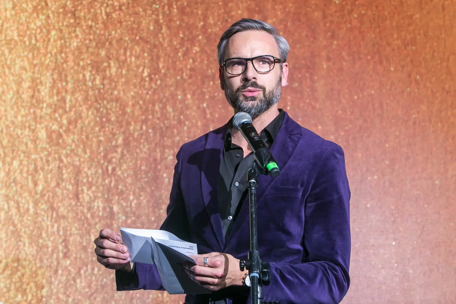 Mężczyzna w fioletowej aksamitnej marynarce otwiera kopertę z werdyktem przy mikrofonie — Gala DMR XI (2022)