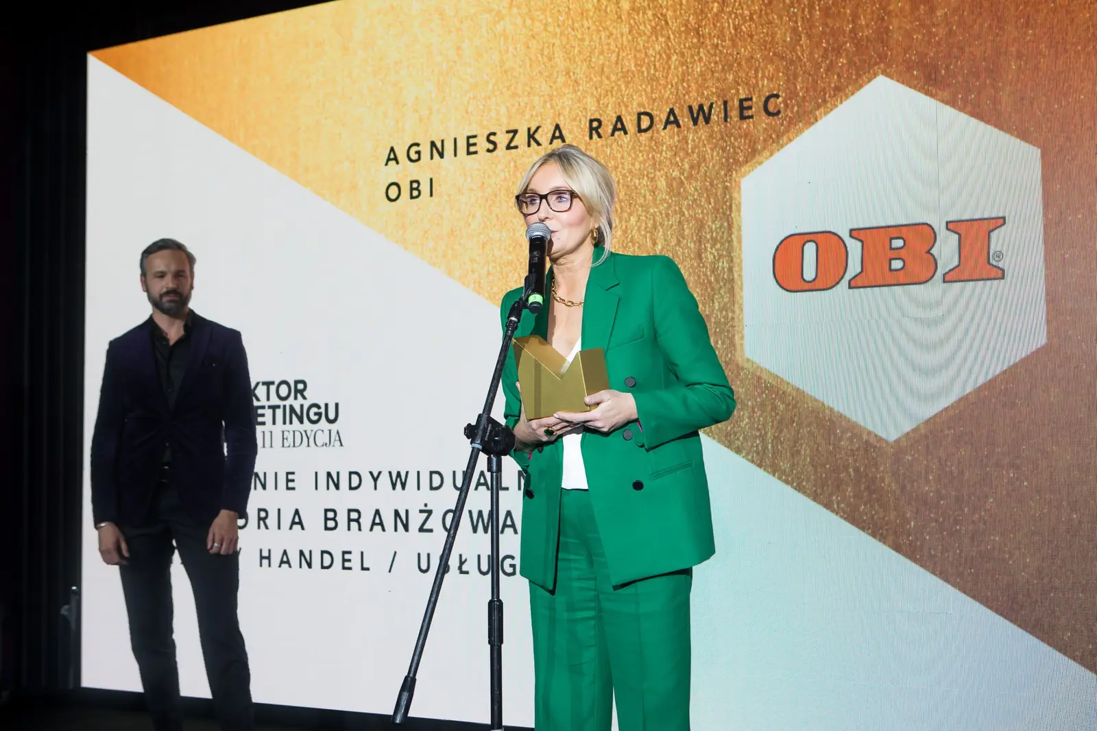 Szeroki kadr: laureatka w zielonym garniturze przemawia w kategorii handel i usługi — Gala DMR XI (2022)